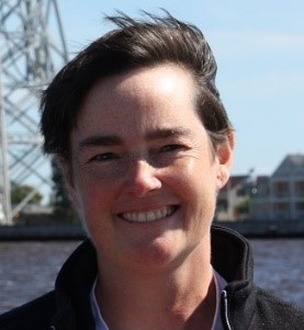 Photo of Amy Jo Swing, head and neck of a woman with short, dark brown hair, wearing collared jacket. She is smiling with water and a part of a bridge in the background.