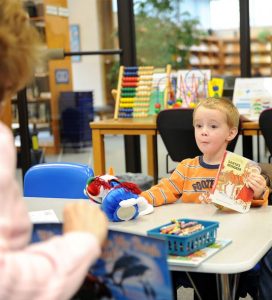 Young boy sitting at table with a teacher. There are books, crayons and toys on the table.