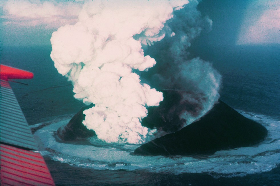 Photo of Surtsey while it was erupting. It shows a volcano rising out of the ocean with a large cloud of ash and steam coming out of the top.