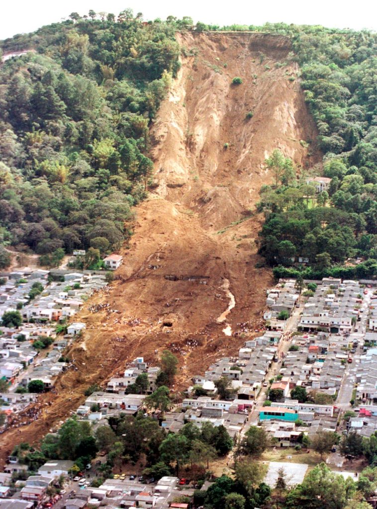 Photograph of a landslide with a large hill in the background and a town in the foreground. The middle portion of the hill has slid down to cover houses.