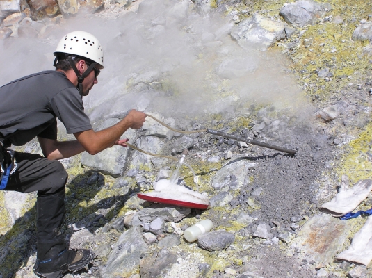 Photograph of a geologist collecting a gas sample by inserting a tube into a fumarole and collecting gas into a flask.